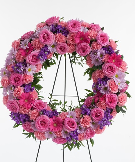 Wreath display featuring a mix of lush pink and lavender flowers including roses, carnations, and daisy's displayed on an easel. 