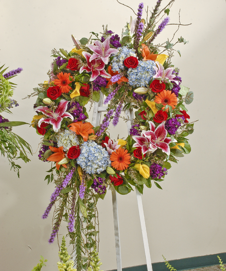 Brightly colored wreath displayed on an easel featuring red roses, lilies, hydrangea and other accent flowers. Approximate size of the standard design is 28