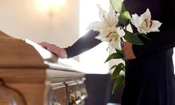 A person in black attire holds white lilies, placing a hand on a wooden casket in a softly lit room, suggesting a somber or memorial setting.